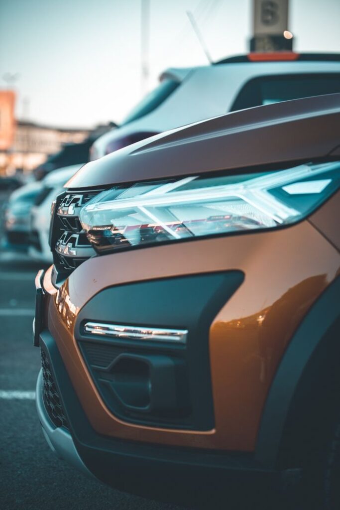 Vertical shot of a car's front, focusing on the illuminated headlight and grill in a parking lot.